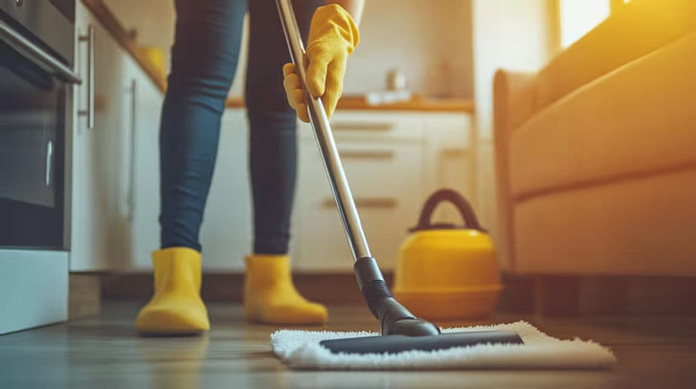 Person cleaning kitchen floor with mop and yellow gloves, warm lighting in background.
